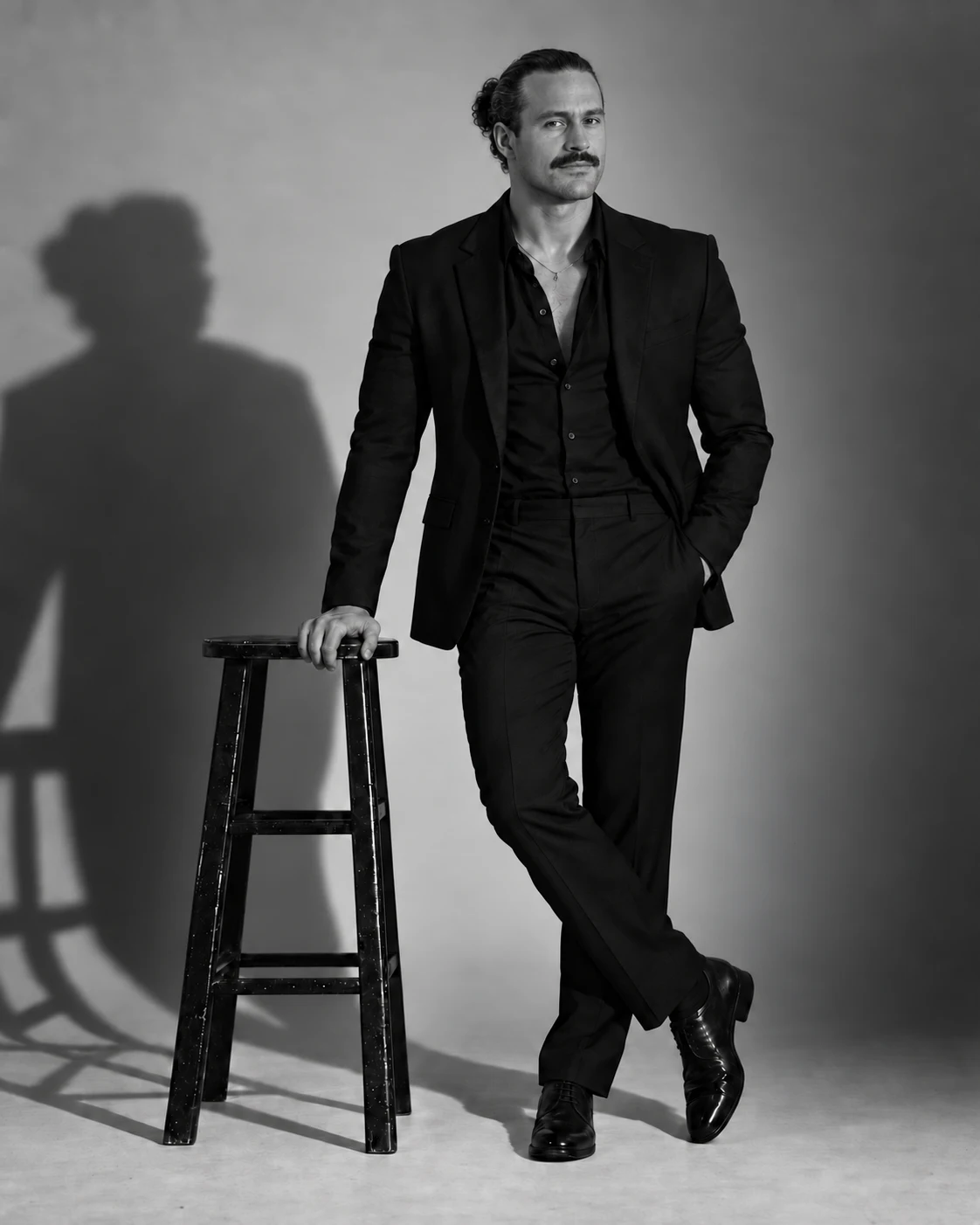 Alex de pie con traje negro junto a un taburete